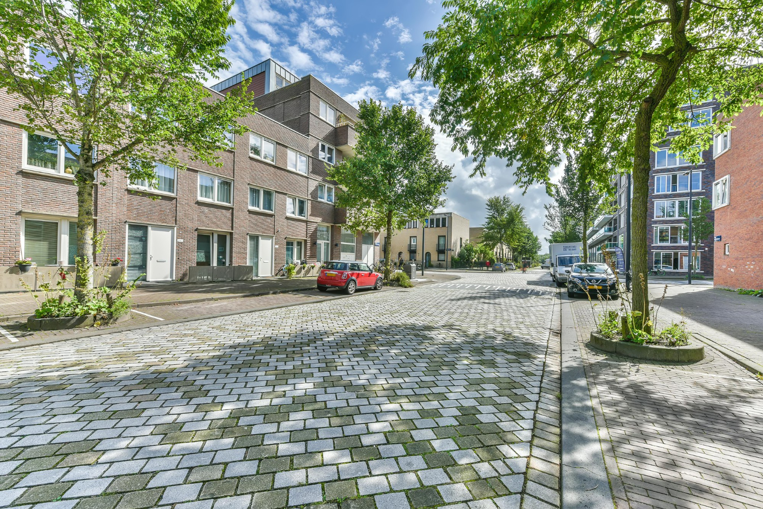 Ulica w spokojnej dzielnicy z niską zabudową mieszkalną z cegły, drzewami rosnącymi wzdłuż chodnika i zaparkowanymi samochodami, w tym czerwonym autem po lewej stronie. Kostka brukowa odbija światło słoneczne, a nad ulicą widoczne jest niebo z lekkim zachmurzeniem.
