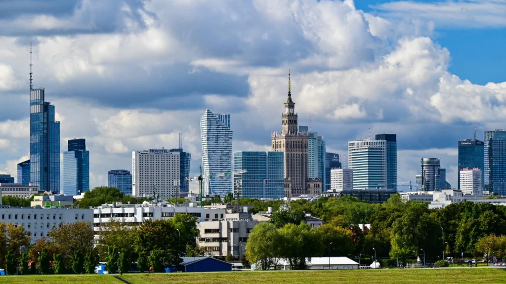 Panorama Warszawy z nowoczesnymi wieżowcami i Pałacem Kultury i Nauki na tle nieba z gęstymi chmurami, widziana z perspektywy zielonego terenu.